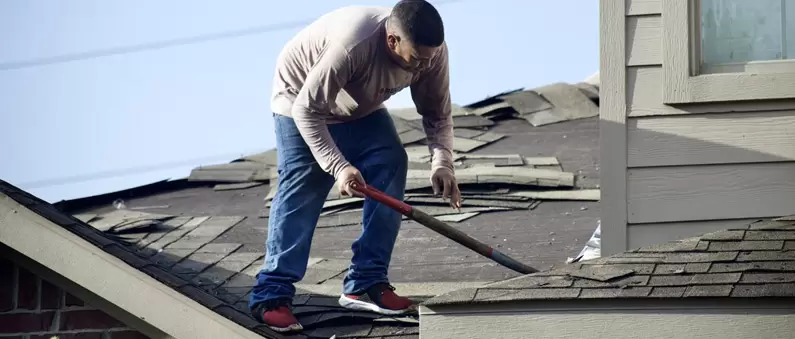 A man uses a shovel to remove tiles during a roof replacement